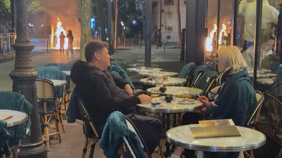 Keep calm and carry on: Unfazed French diners enjoy a drink at Parisian cafe during riots as fire burns in the background